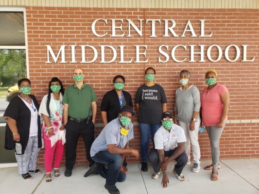 People in face masks pose in front of brick wall with large letters reading "Central Middle School"